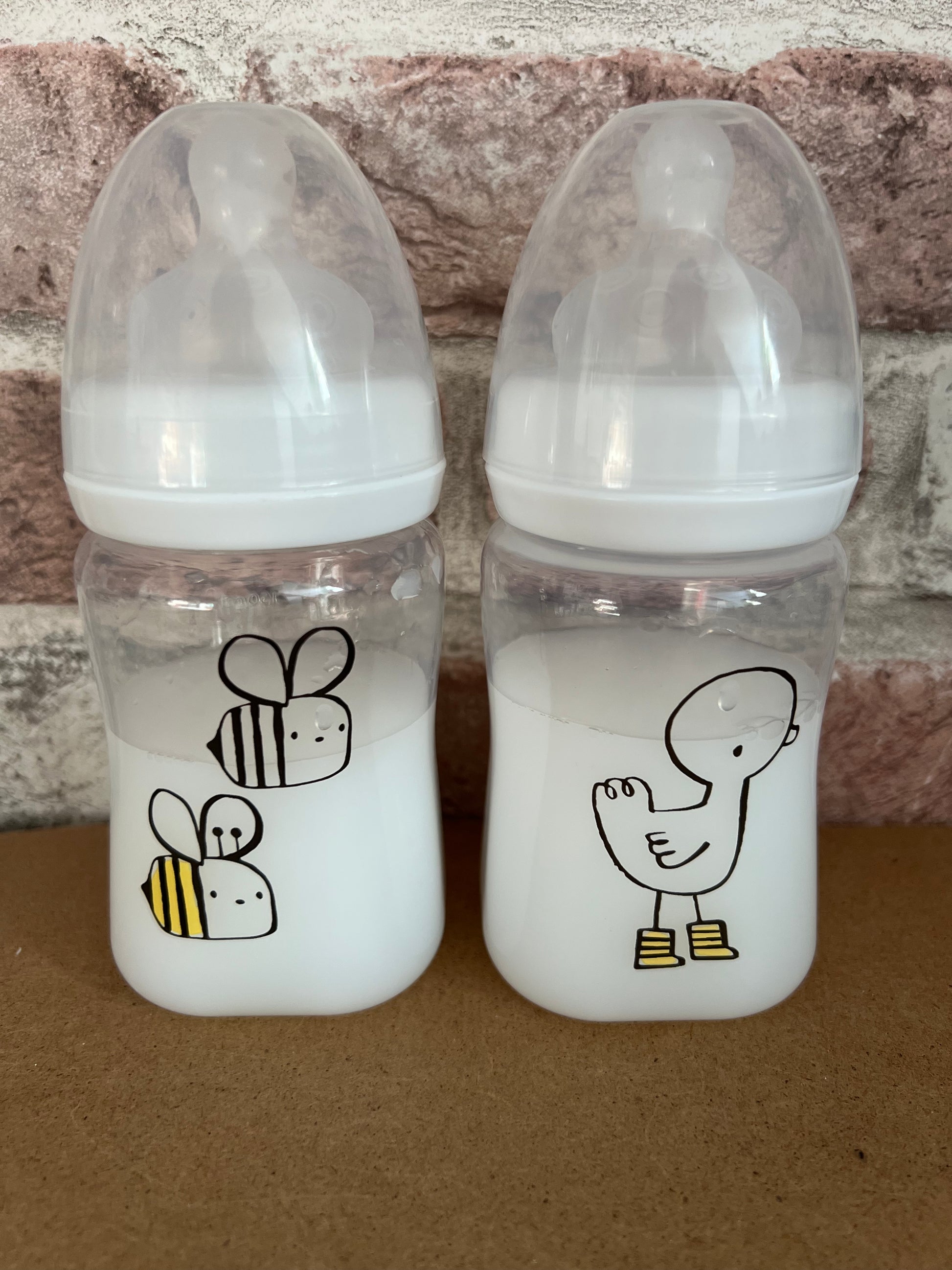 Two baby bottles with cartoon bee and duck designs on a stone background - willow tree reborns UK