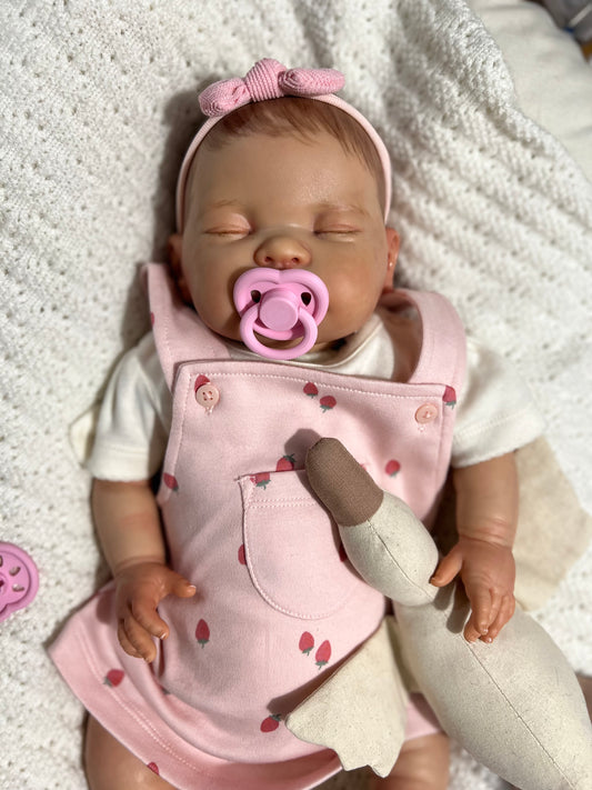Reborn baby doll wearing a pink outfit with a sugar snap pacifier on a soft surface